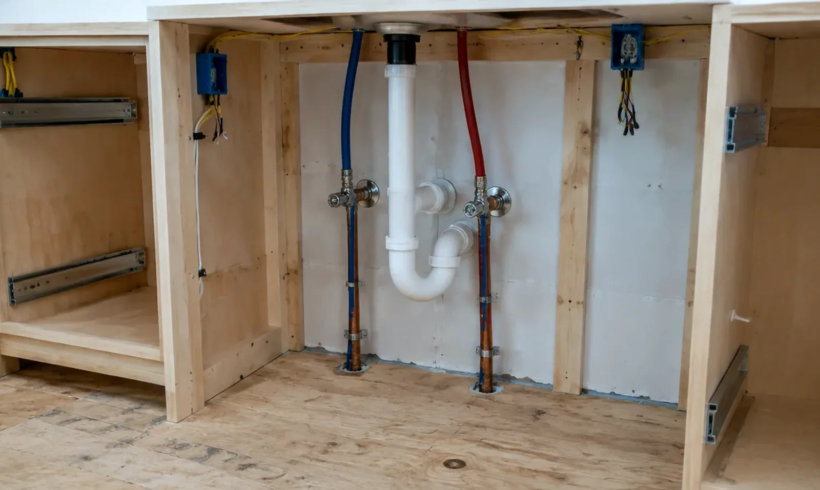 Illustrative: open sink base with plumbing supply lines, drain trap, and electrical boxes roughed in before cabinets and countertops go back in — illustrative only, not a real Board of Project Stewardship job photo